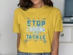 A woman wearing a yellow t-shirt with the phrase "STOP LOOKING AT MY TACKLE" in blue text, standing indoors.