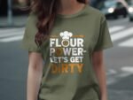 A woman in an olive green t-shirt with the text "FLOUR POWER- LET'S GET DIRTY," featuring a chef's hat and whisk graphic.