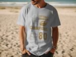 A man in a light blue t-shirt with gold football-themed graphics stands on a beach.
