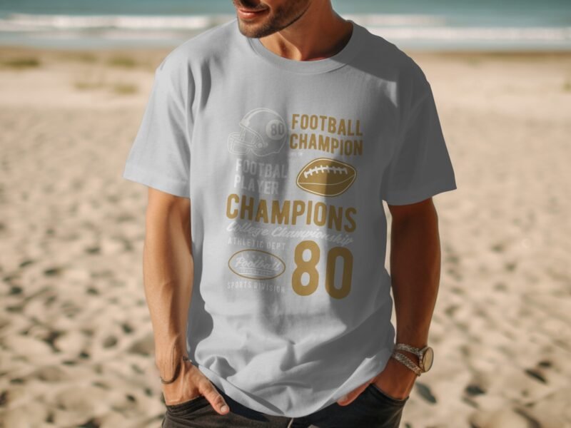 A man in a light blue t-shirt with gold football-themed graphics stands on a beach.