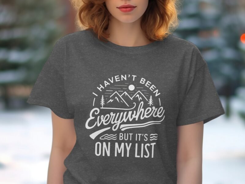A person wearing a gray t-shirt with the quote "I haven't been everywhere but it's on my list," and mountain graphics.