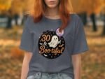 A woman in a gray t-shirt with a ghost graphic reads "30 & Bootiful," surrounded by autumn trees.
