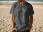 A man in a dark gray t-shirt with a motorcycle graphic stands on a sandy beach with ocean waves in the background.