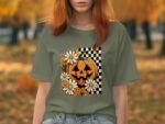 A woman wears a green t-shirt featuring a pumpkin design with flowers and a checkerboard background, set in an autumn scene.
