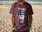 A man wearing a purple t-shirt featuring a vintage van graphic and beach-themed text, standing on a sandy beach.