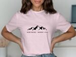 A woman in a pink t-shirt with "Hike More - Worry Less" and mountain graphic, smiling with hands on her hips.