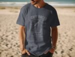 A man wearing a navy t-shirt with a vintage motorcycle design stands on a sandy beach with ocean waves in the background.