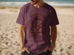 A man wearing a purple t-shirt with butcher-related graphics stands on a beach with ocean waves in the background.
