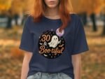 A woman wears a blue t-shirt with a ghost graphic and the text "30 & Bootiful" against a fall backdrop.