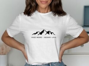 Woman wearing a white t-shirt with a mountain graphic and the text "HIKE MORE - WORRY LESS." Minimalist background.