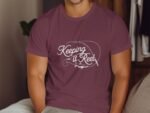 A man wears a maroon T-shirt with the phrase "Keeping it Reel" and fishing imagery, set against a neutral background.