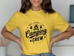 A woman wearing a yellow t-shirt with a black "Camping Crew" graphic, posing playfully with hands on hips.