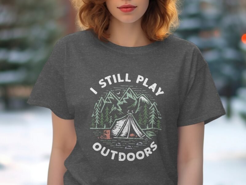 Woman wearing a gray t-shirt with "I STILL PLAY OUTDOORS" text and a camping graphic featuring mountains and a tent.