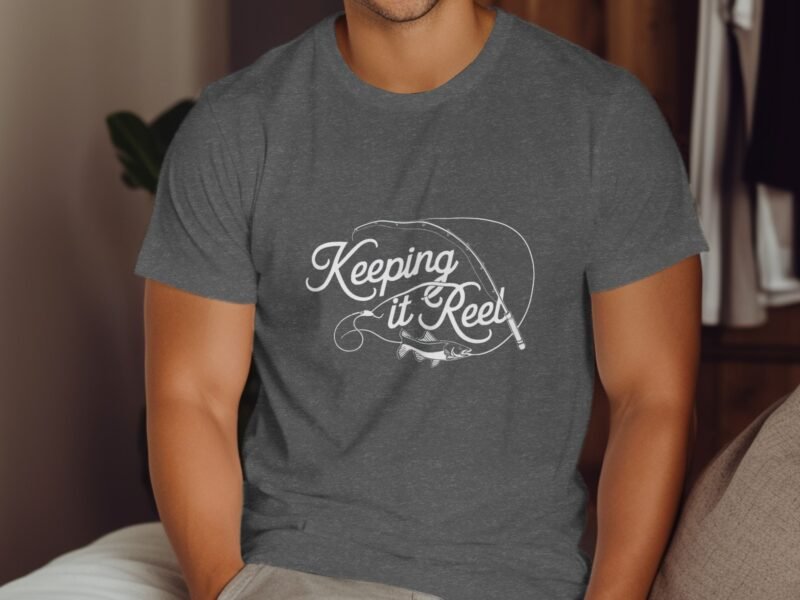 A man wearing a gray t-shirt with the text "Keeping it Reel" and a fishing theme graphic, sitting indoors.