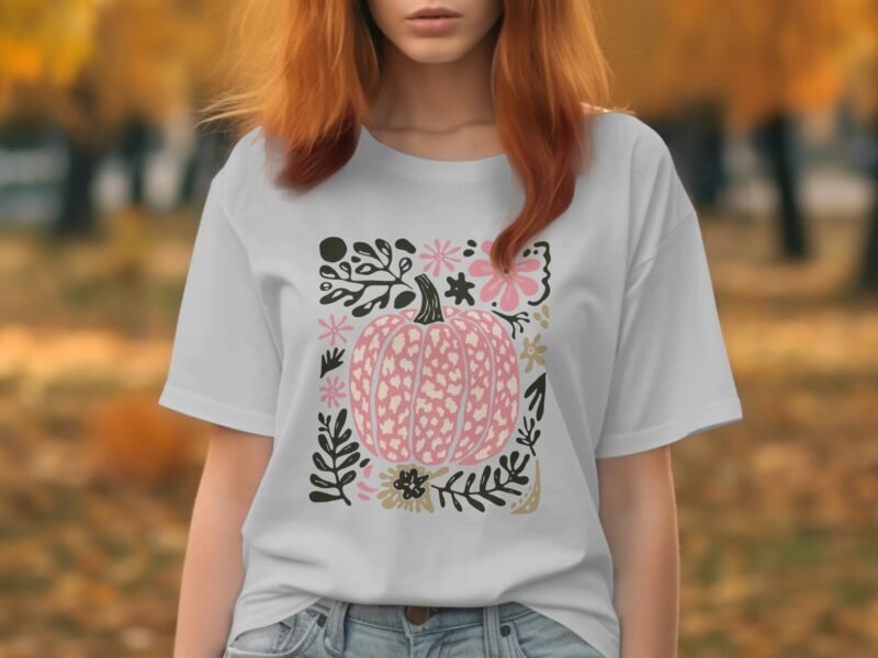 A woman with long, red hair wears a gray t-shirt featuring a pink pumpkin and floral design, with an autumn backdrop.