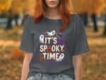 A woman in a gray t-shirt with a spooky design stands in a fall landscape with orange leaves.