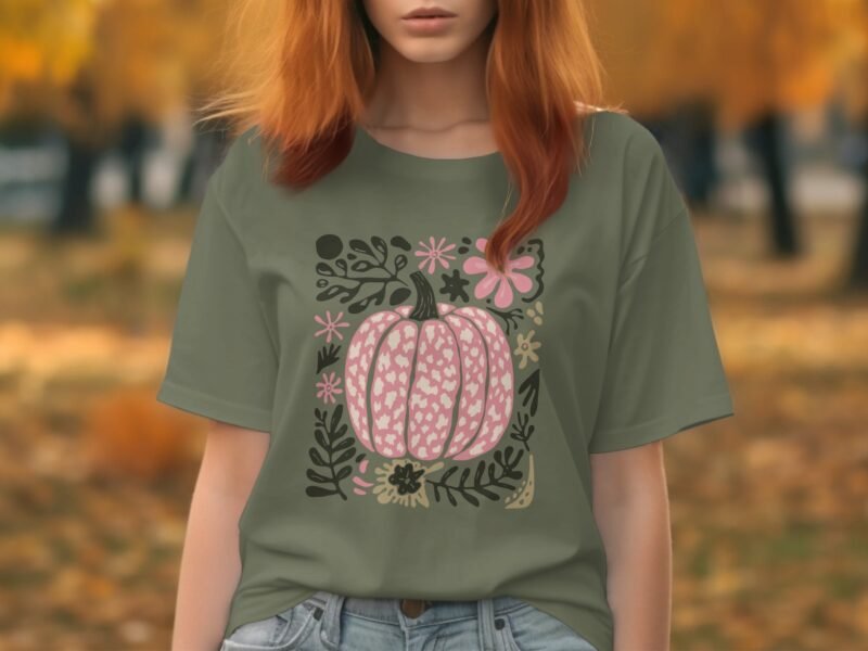 A person wearing a green t-shirt with a pumpkin and floral design, surrounded by autumn foliage.