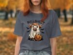 Woman in a gray T-shirt featuring a cartoon cow in a ghost costume with a pumpkin, set against a fall background.