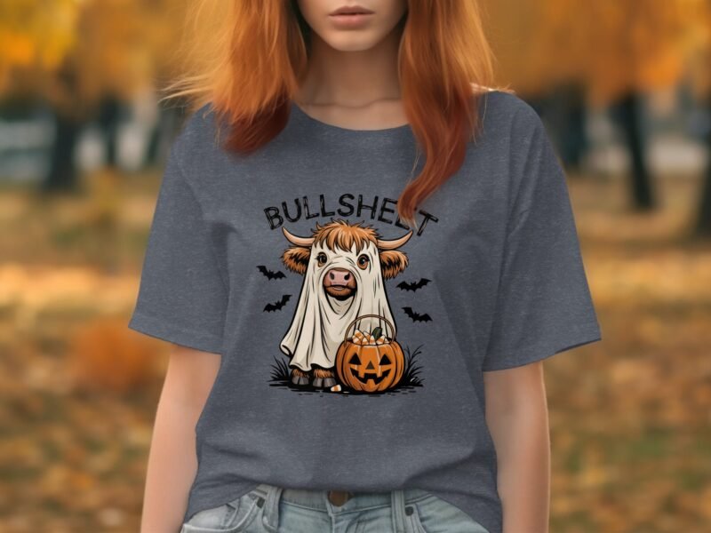 Woman in a gray T-shirt featuring a cartoon cow in a ghost costume with a pumpkin, set against a fall background.