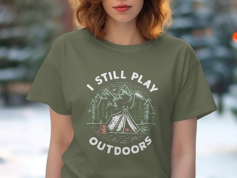 Woman wearing an olive green t-shirt with "I STILL PLAY OUTDOORS" text and a camping scene graphic.