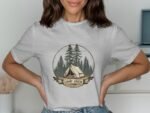 Woman wearing a grey t-shirt with a "Camp Crew" graphic featuring a tent and pine trees. Interior setting visible.
