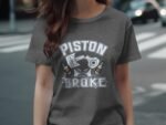 A woman wearing a gray t-shirt featuring the text "PISTON BROKE" and illustrations of a piston and bottles.