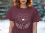 A woman with wavy hair wears a maroon T-shirt featuring a white mountain and sun design with rays and trees.