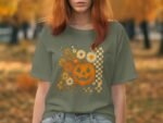 A woman with reddish hair wears an olive green t-shirt featuring a smiling pumpkin and floral design, set in an autumn background.