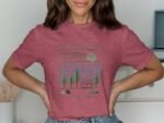 A woman wearing a pink graphic t-shirt featuring a camper, trees, and a sun in a scenic landscape design.