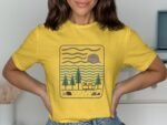 A woman in a yellow t-shirt with a camping scene illustration featuring trees, a trailer, sun, and waves.