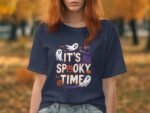 A woman with red hair wearing a navy t-shirt that says, "It's Spooky Time," surrounded by autumn leaves.