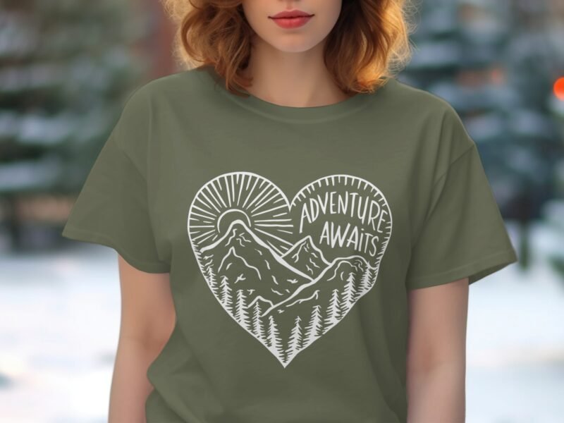 A woman wearing an olive green t-shirt with a heart design featuring mountains, trees, and the phrase "Adventure Awaits."