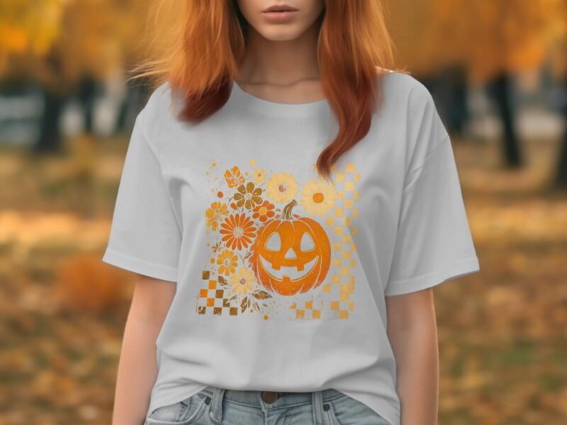 Woman in a light gray t-shirt featuring a smiling pumpkin graphic and flowers, outdoors with autumn foliage in the background.