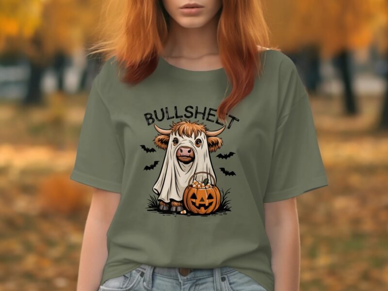 A person in a forest wears an olive green shirt featuring a cartoonish cow in a ghost costume holding a pumpkin.