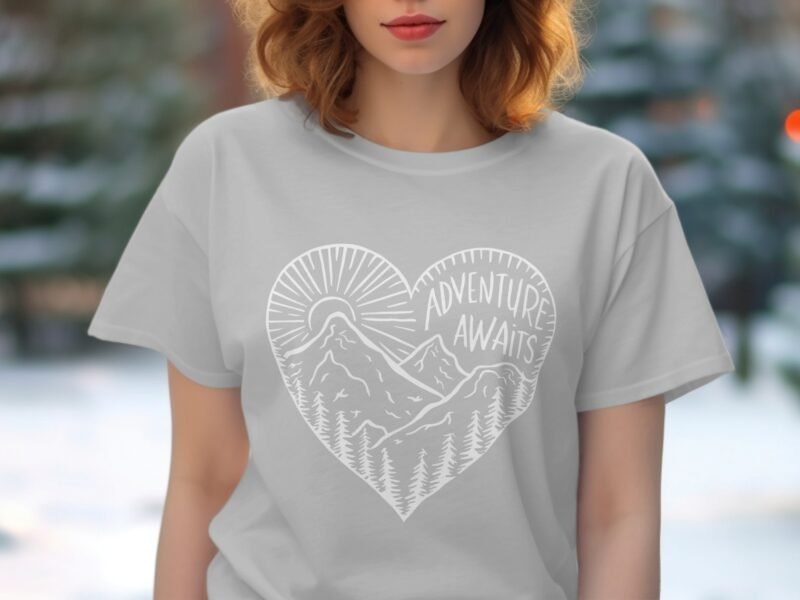 A woman wearing a light gray t-shirt with a heart design featuring mountains and the phrase "Adventure Awaits."