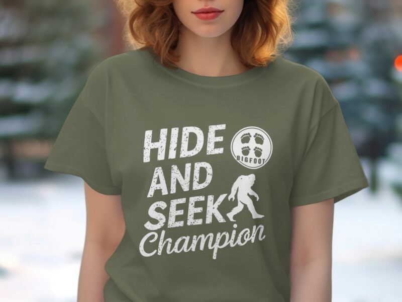 Woman in an olive green t-shirt with "HIDE AND SEEK Champion" and a Bigfoot graphic, outdoors with blurred trees in background.