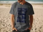 Man in a gray t-shirt with a vintage van graphic and text about adventure and monotony, standing on a beach.