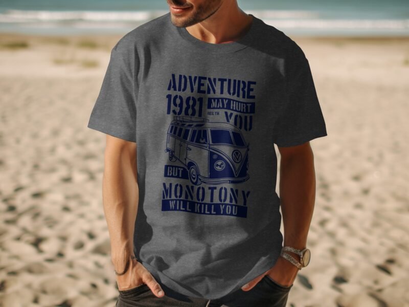 Man in a gray t-shirt with a vintage van graphic and text about adventure and monotony, standing on a beach.