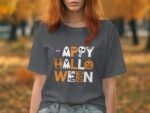 A woman with reddish hair wears a gray t-shirt featuring "Happy Halloween" in festive, colorful letters. Autumn leaves background.