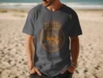 A man wearing a dark gray t-shirt with a vintage car graphic stands on a sandy beach with ocean waves in the background.