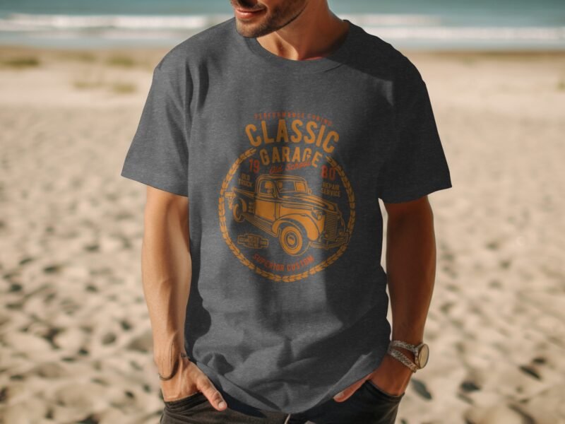A man wearing a dark gray t-shirt with a vintage car graphic stands on a sandy beach with ocean waves in the background.