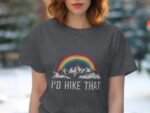 A woman in a gray t-shirt featuring a rainbow over mountains and the text "I'd Hike That." Background with green trees.