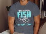 A man wears a gray t-shirt with the phrase "I CATCH MY FISH IN REEL TIME" and a graphic of a fishing scene.
