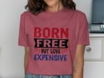 A woman wears a maroon t-shirt with the text "BORN FREE BUT LOVE EXPENSIVE" in bold, colorful fonts.