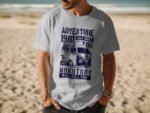 A man in a light blue t-shirt with a vintage van graphic and text about adventure and monotony, standing on a beach.