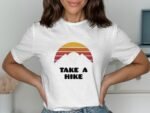 A woman in a white t-shirt with a sunset and mountains graphic, featuring the text "TAKE A HIKE."