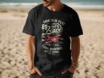 Man wearing a black t-shirt featuring a vintage airplane graphic and text about aviation adventures, on a beach.