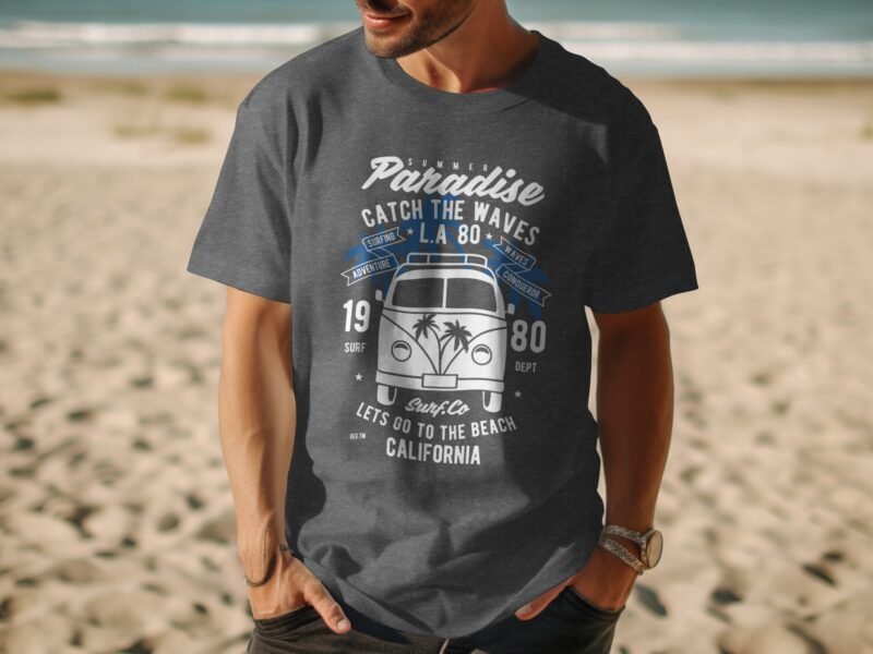 A man wearing a dark gray t-shirt with surf-themed graphics, standing on a beach with ocean waves in the background.