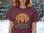 A woman in a maroon t-shirt with a graphic of trees and sunset in yellow and blue, set against a blurred background.
