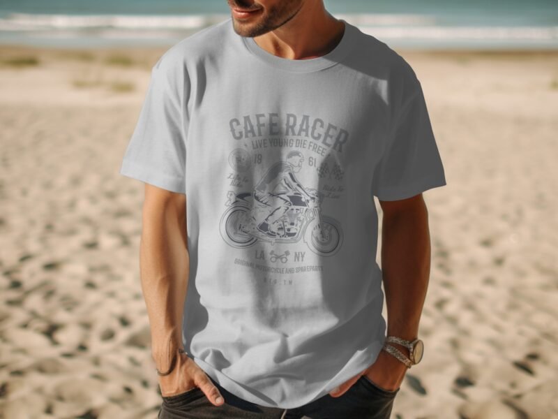 A man in a light gray t-shirt with a motorcycle graphic stands on a beach, with ocean and sand in the background.